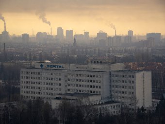 Fotografia wielkiego betonowego szpitala z niebieskim napisem SZPITAL na tle zamglonej, smogowej panoramy miasta z dymiącymi kominami