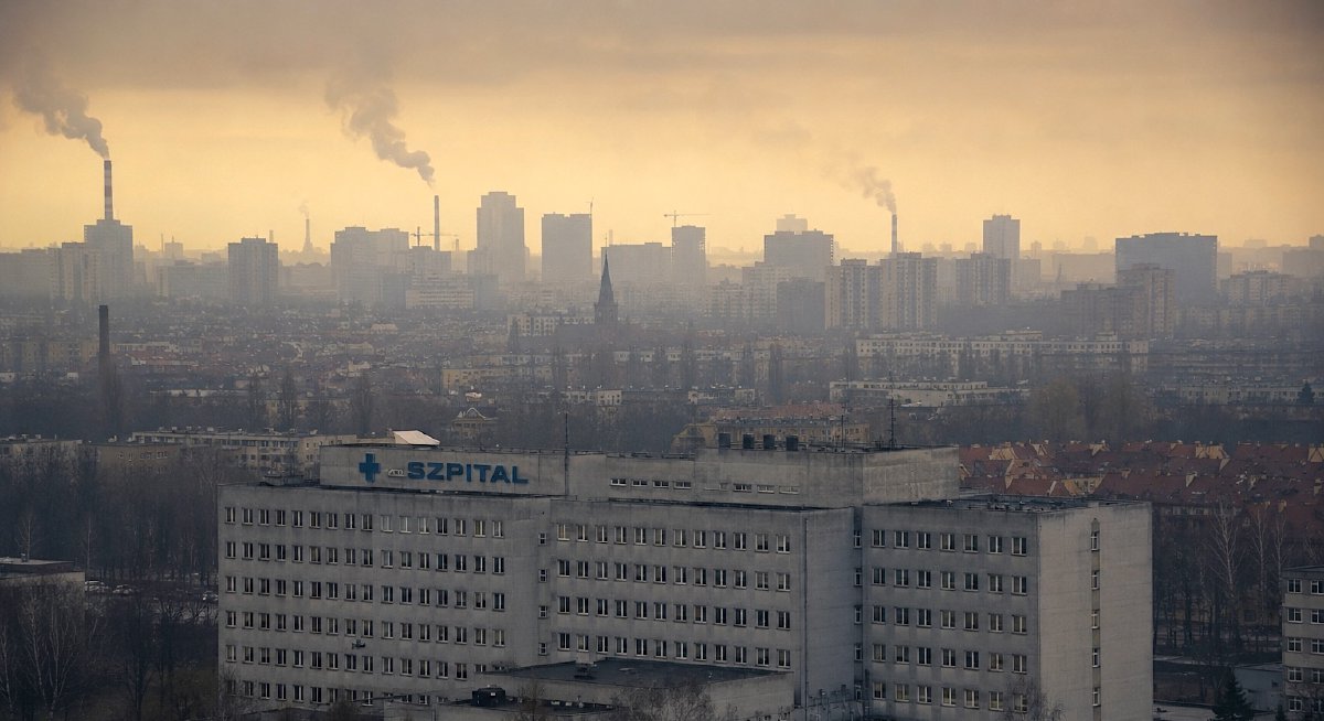 Fotografia wielkiego betonowego szpitala z niebieskim napisem SZPITAL na tle zamglonej, smogowej panoramy miasta z dymiącymi kominami
